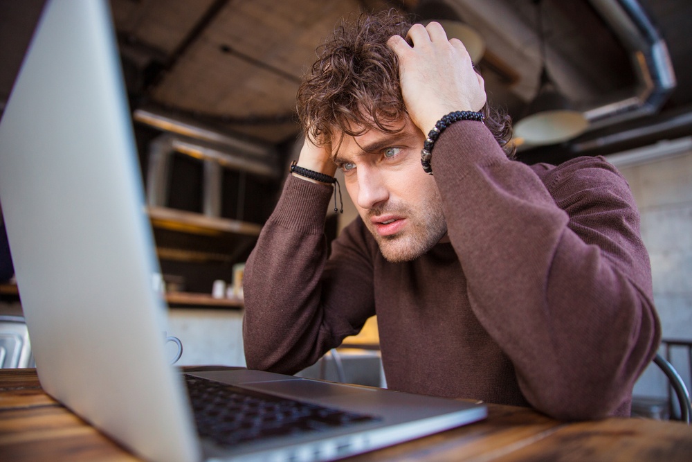 Stressful upset desperate handsome curly man in brown sweetshirt working using laptop and having headache